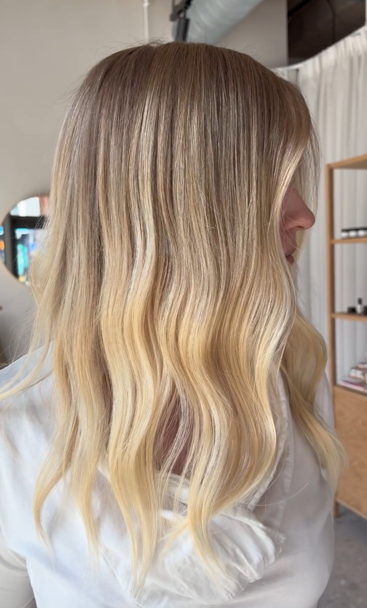 Long, wavy blonde hair with a balayage gradient, shown from the back on a person wearing a white top in a salon setting.
