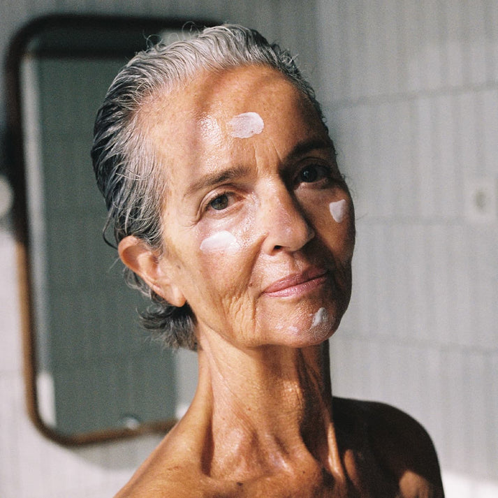 A close up image of a woman with her hair brushed back off her face and dabs of cream on her cheeks forehead and chin