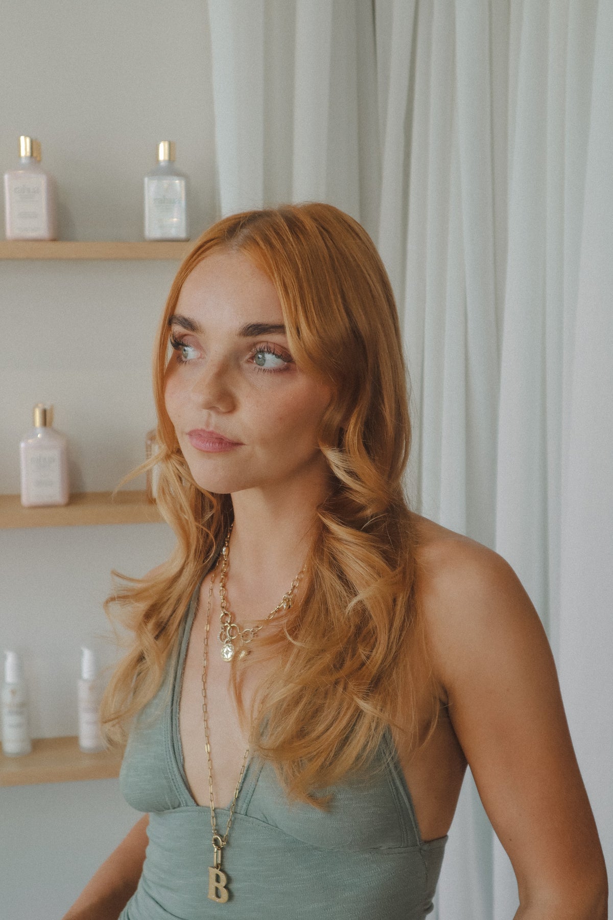A woman with long, wavy strawberry blonde hair Glasshouse salon Bespoke hair treatment and blowdry is shown from the chest up, wearing a sage green halter top and layered gold necklaces. She is standing in a brightly lit room with white curtains and shelves containing skincare bottles in the background.