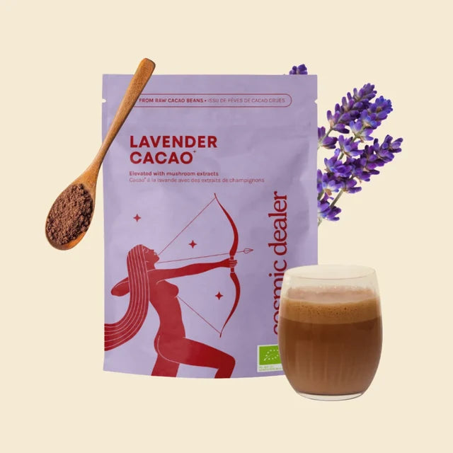 A stand-up pouch of Cosmic Dealer Lavender Cacao powder, shown with a wooden spoon of the dark brown cacao powder, a glass of the prepared brown drink, and purple lavender flowers on a light beige background.