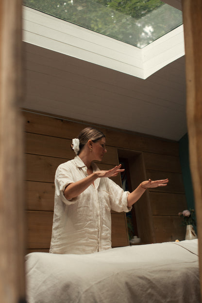 Olivia Crighton in a white linen shirt stands in a sunlit room with wood-paneled walls and a skylight, energy reiki healing 