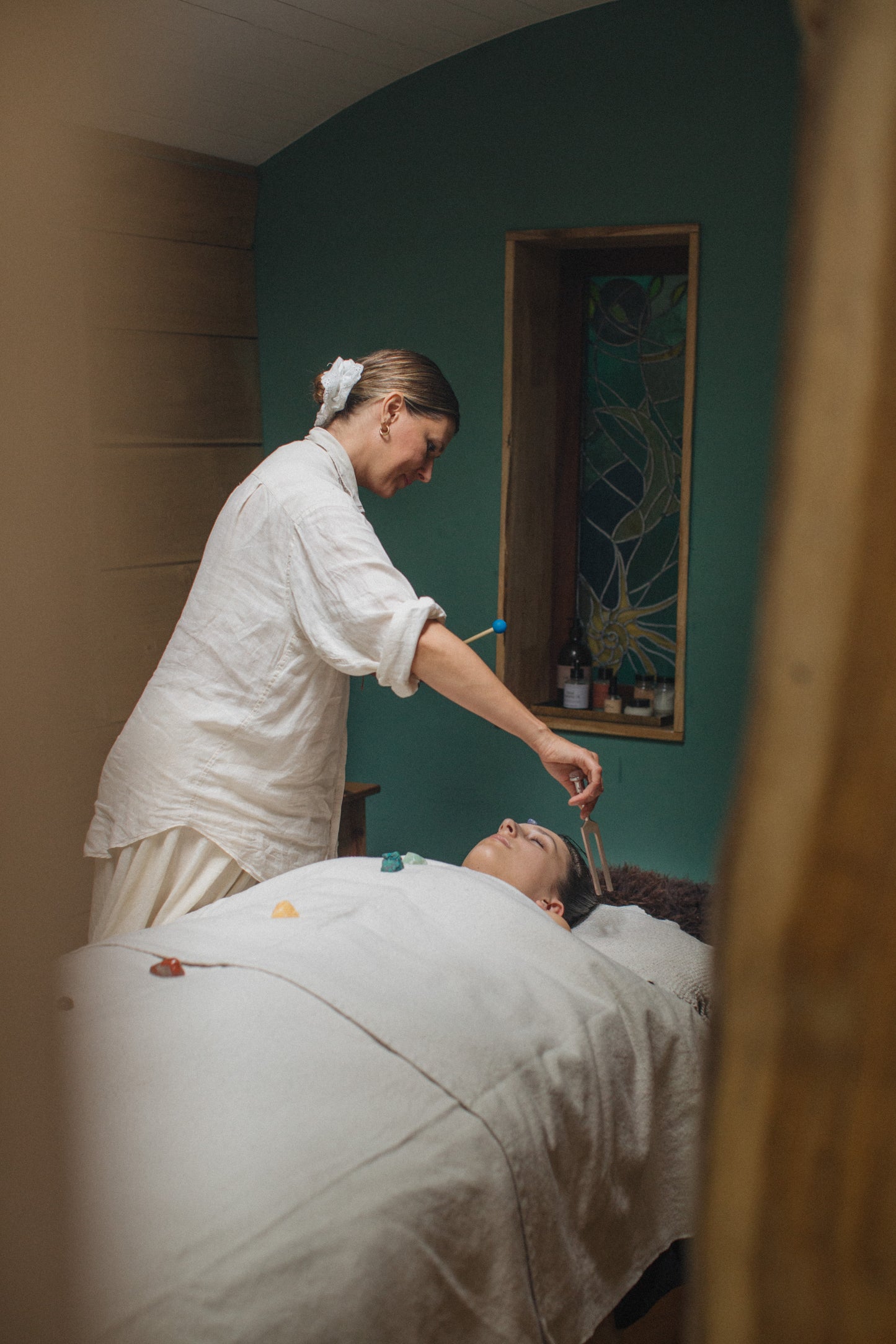 A lifestyle scene of a wellness practitioner in a white linen uniform administering a facial treatment to a client lying down on a treatment bed in a spa room with teal walls.