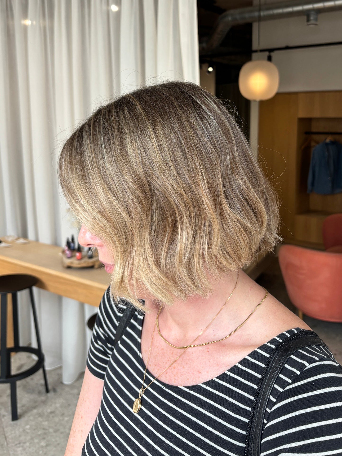 A woman shown from the side profile wearing a short, wavy blonde bob haircut. The hair features a balayage style with warm golden-blonde highlights and darker roots.