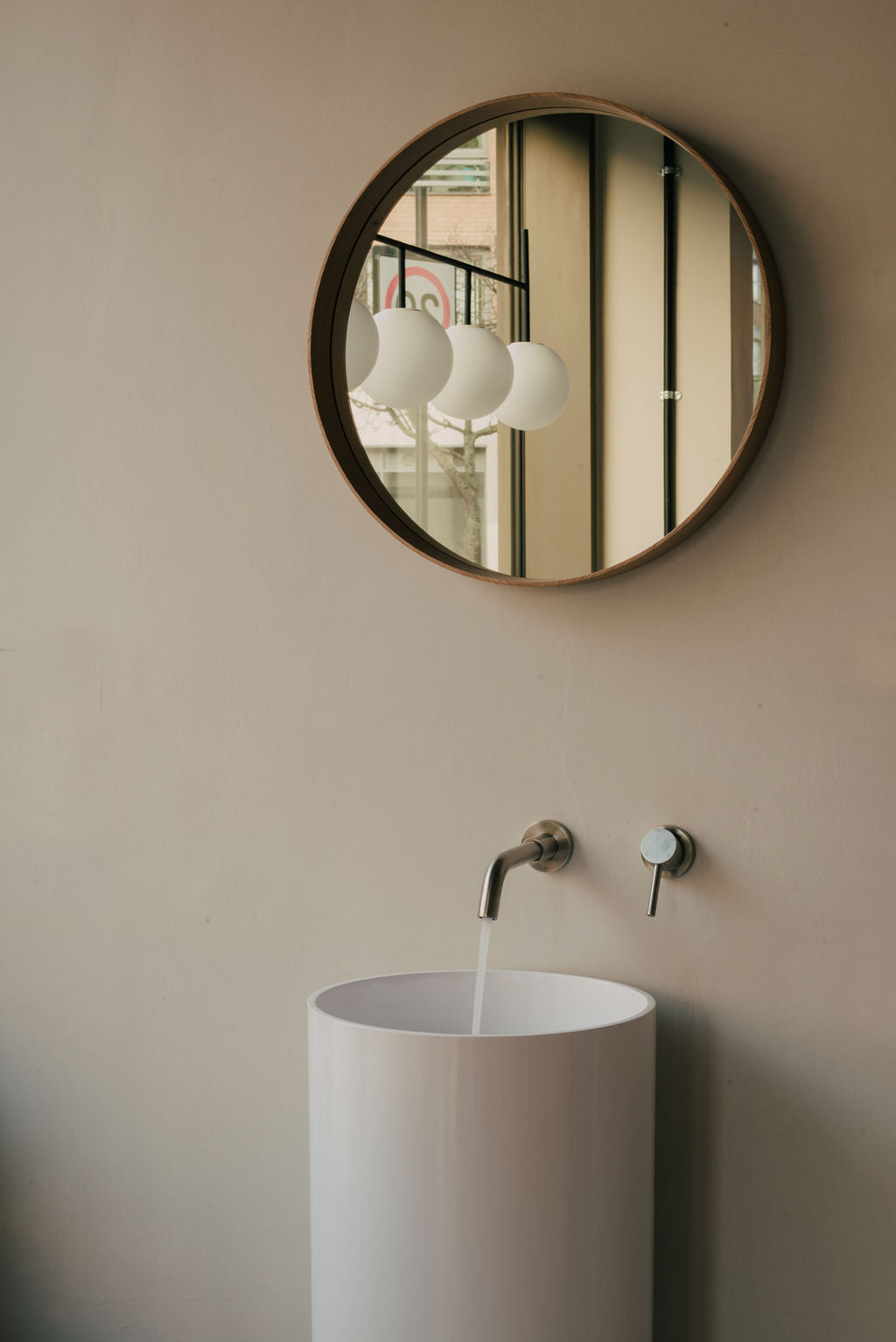 Round mirror on a beige wall above a white sink with running water at Glasshouse Salon