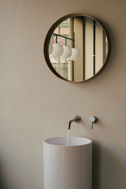 Round mirror on a beige wall above a white sink with running water at Glasshouse Salon