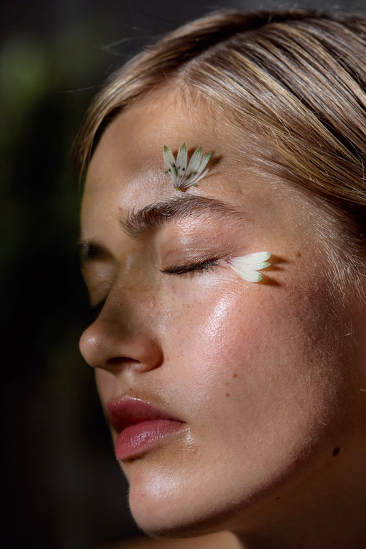 Close-up side profile of a woman with closed eyes, wearing delicate white flower petals on her forehead and cheek, against a dark blurred background.