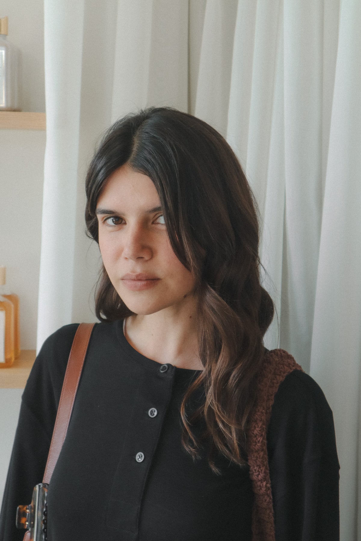A woman with long dark brown hair wearing a black button-up top and a brown textured shoulder bag, standing in front of white curtains at Rahua ritual blowdry event