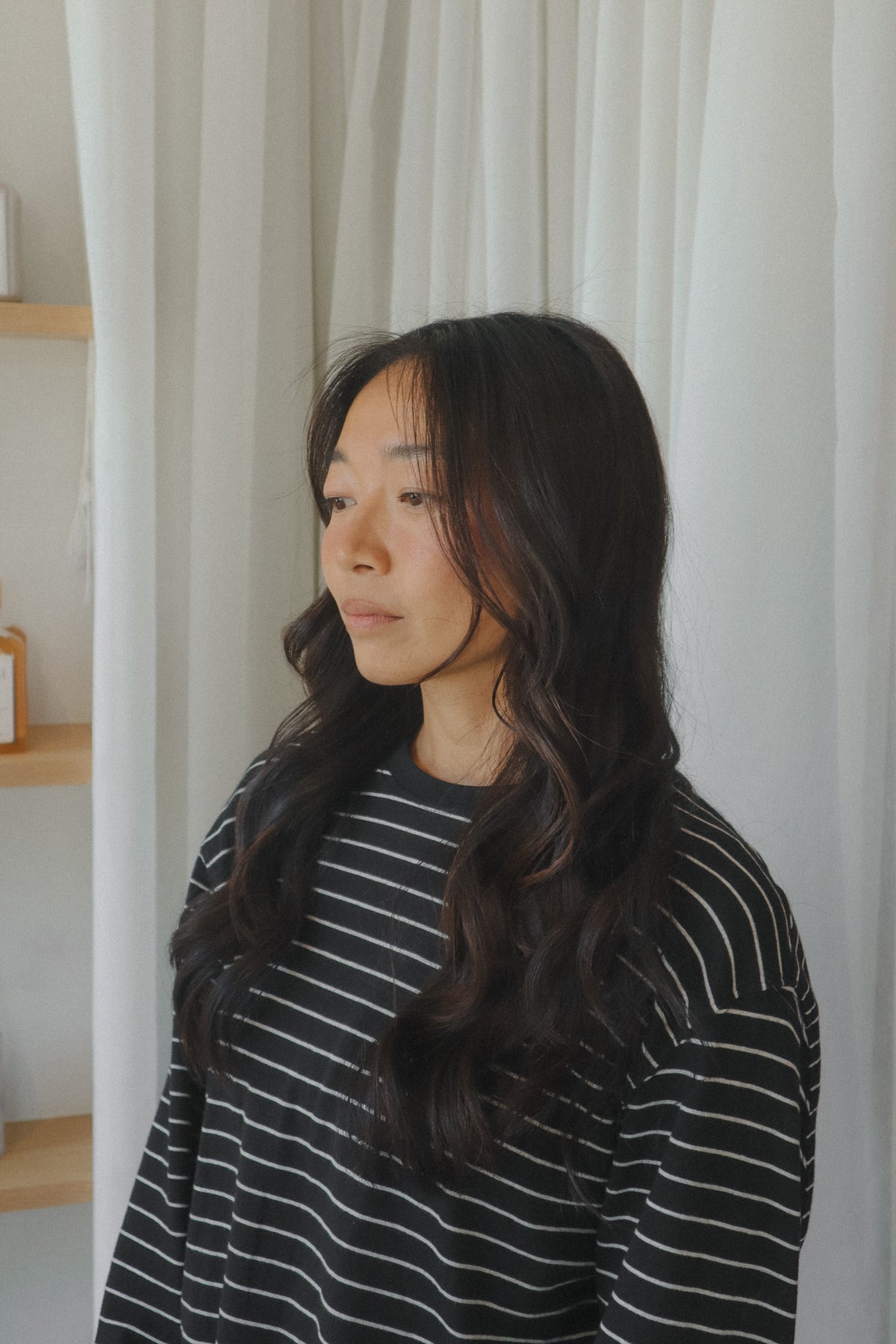 A woman with long, wavy dark hair is shown in profile, wearing a black long-sleeve top with thin white horizontal stripes at Glasshouse Salon 