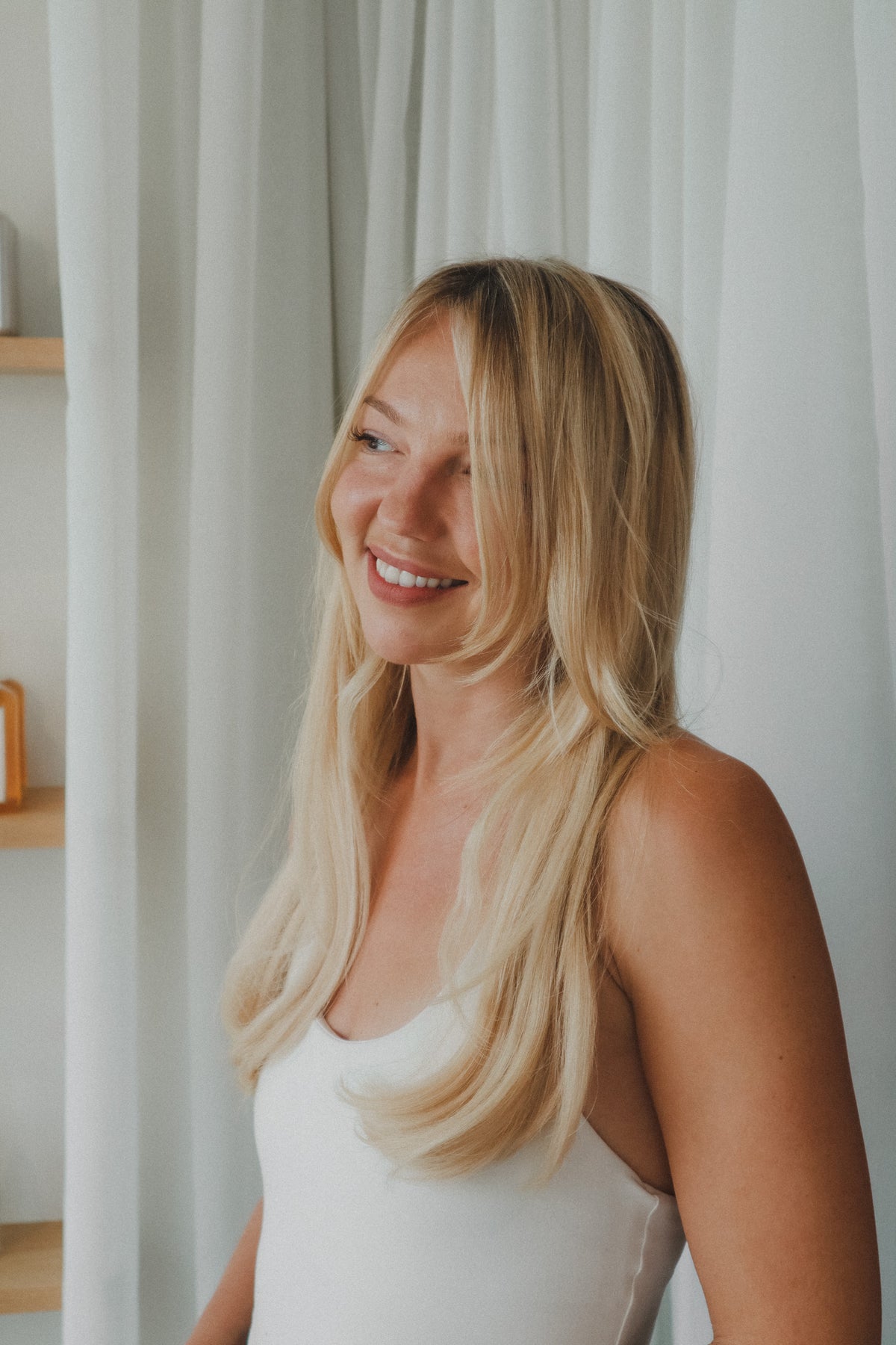 A smiling blonde woman with long wavy hair wearing a white tank top, standing in front of white curtains at Glasshouse Salon