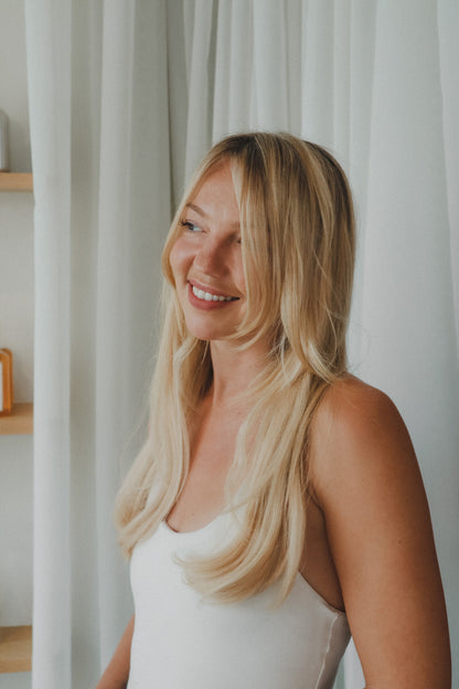 A smiling blonde woman with long wavy hair wearing a white tank top, standing in front of white curtains at Glasshouse Salon