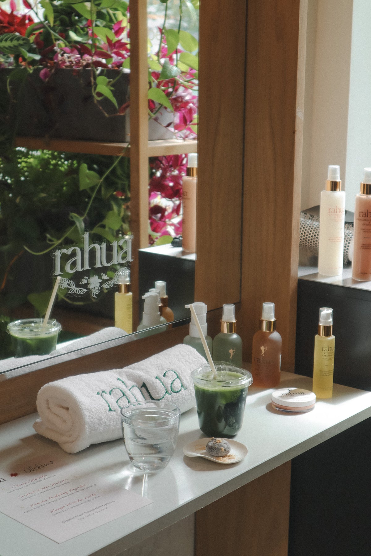 A spa vanity scene with a rolled white towel printed with the brand name 'rahua', a clear glass, a small dish with a spoon, a green drink in a plastic cup, and several bath/body product bottles arranged on a white counter in front of a mirror.