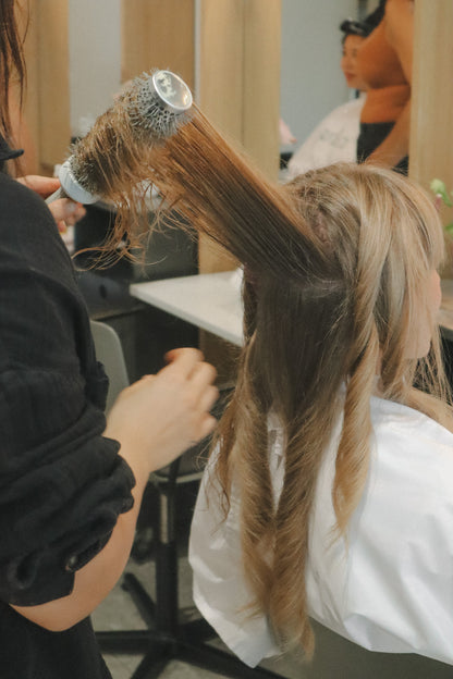 A hairdresser uses a round hair dryer with a brush attachment to style a client's long, light brown hair in a salon setting.