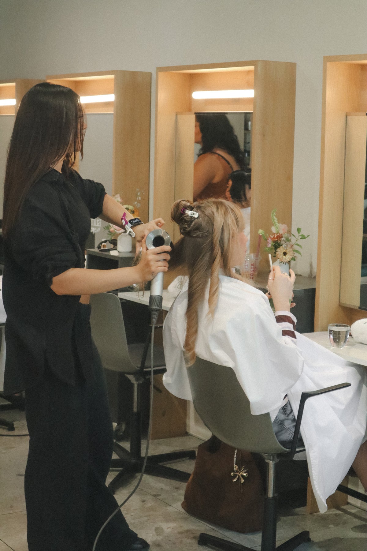 A hairdresser in a black salon uniform styles a client's hair with a curling iron in a modern salon with wooden-framed mirrors and warm lighting.