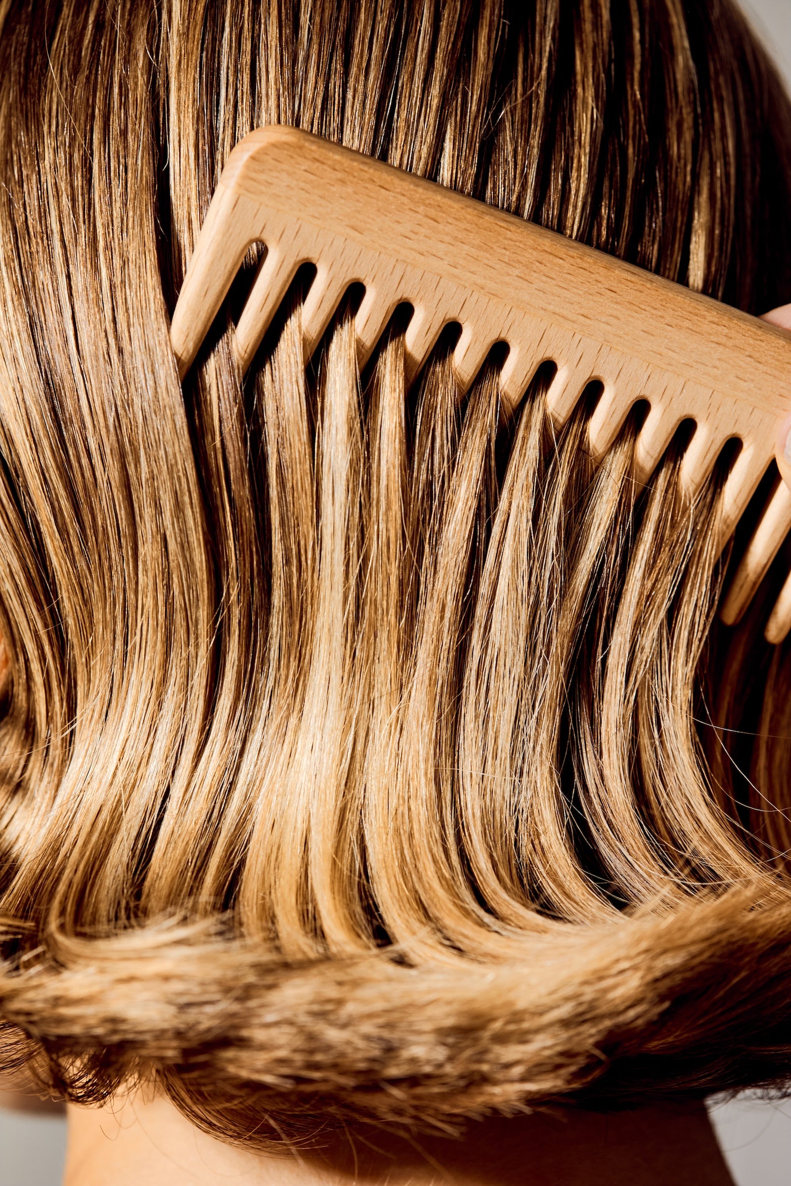 A close-up of a light natural wood comb being used to brush through styled brown hair.