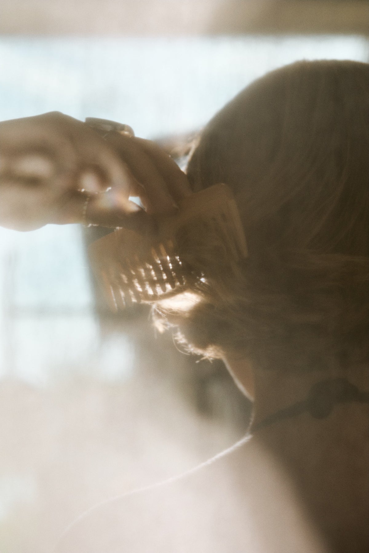 A close-up lifestyle shot of a person with light brown hair using a large, textured wooden hair claw clip. The image is heavily backlit with warm, hazy sunlight, creating a soft, dreamy aesthetic.