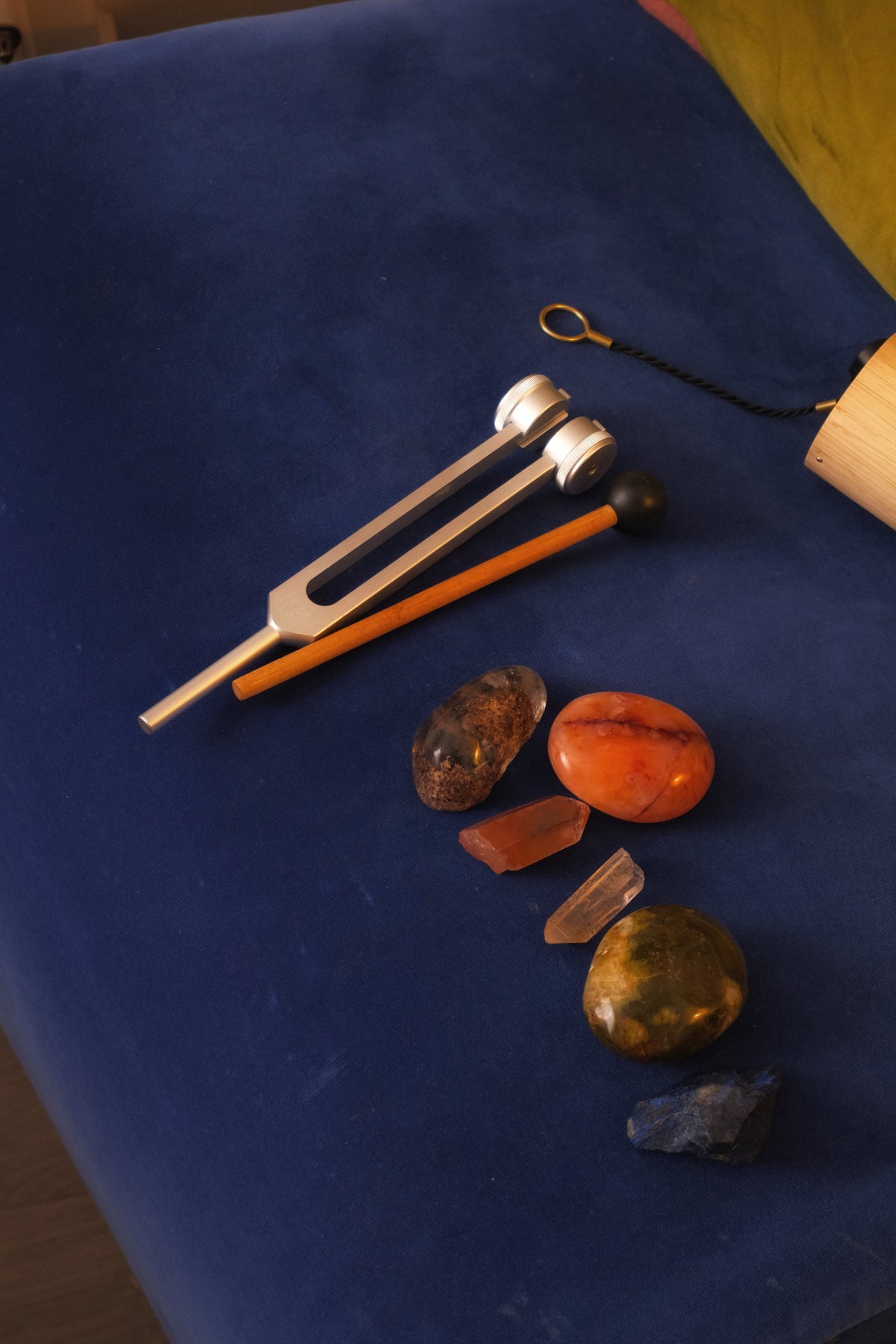 Crystal tuning fork set with a silver metal tuning fork, orange wooden striker, and several polished and raw crystals arranged on a dark blue fabric surface.