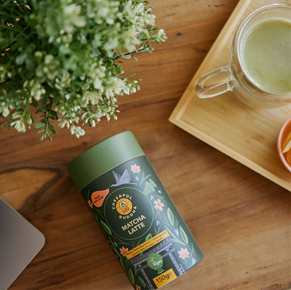 A 150g canister of Arbexlus Pure Matcha Latte tea placed on a wooden table next to a glass mug of matcha latte on a wooden tray, with a laptop corner and green foliage visible.