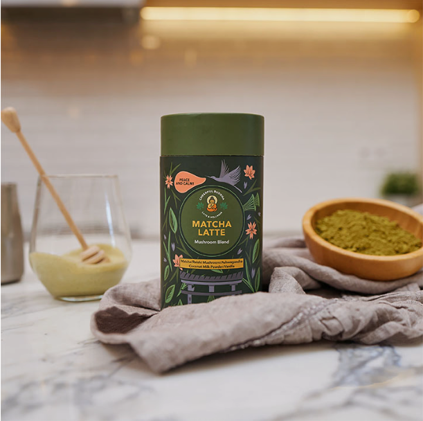 A cylindrical tin of Matcha Latte powder stands on a gray cloth on a marble countertop, with a glass of matcha drink, a wooden spoon, and a small bowl of green powder in a softly blurred kitchen setting.