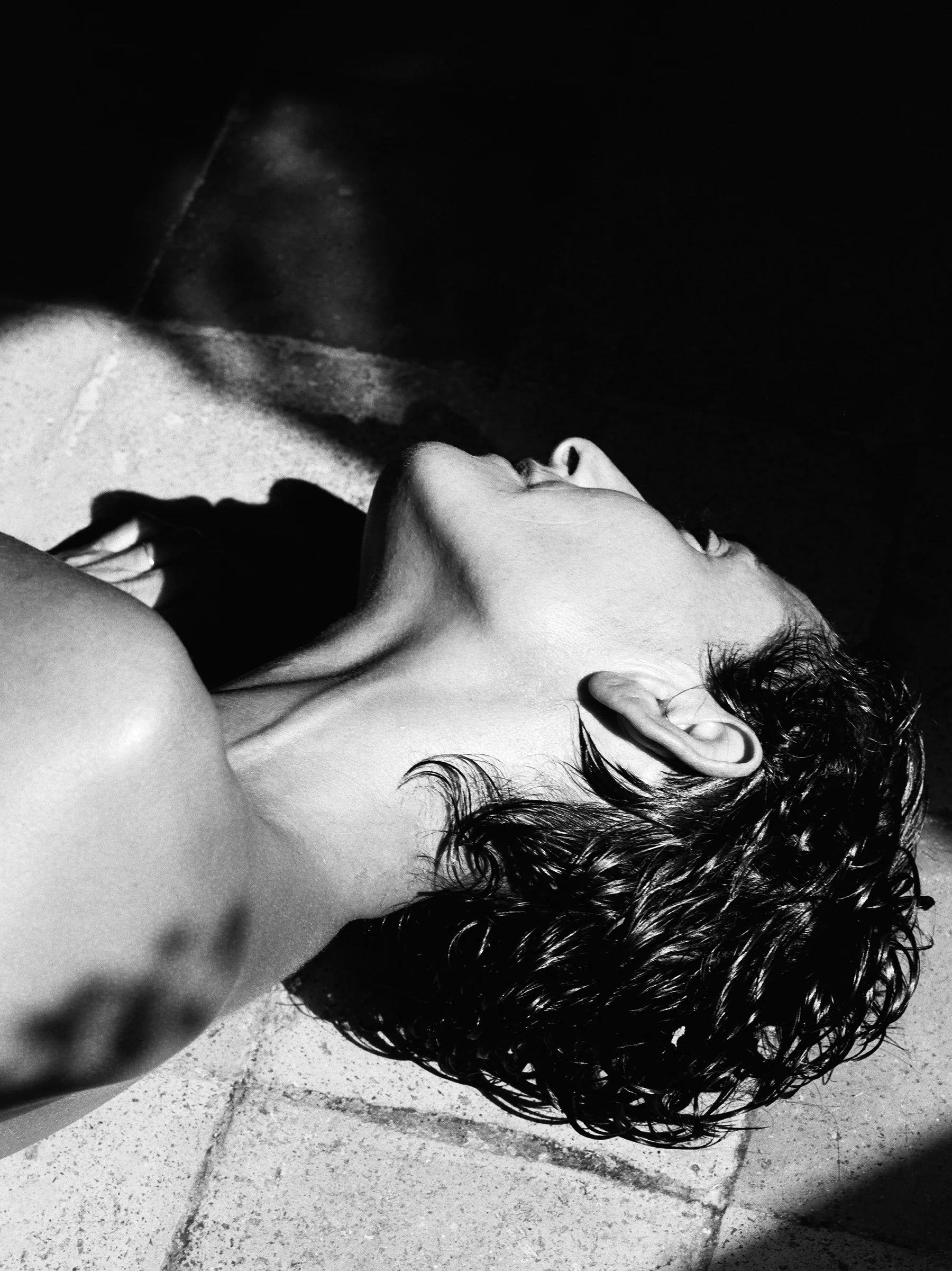 Black and white image of a woman with short hair lying on a tiled floor looking away from the camera