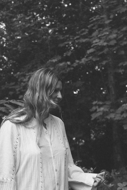 A black and white photograph of an energy healer Olivia Crighton with long hair wearing a loose, long-sleeve button-up shirt with decorative stitching, standing outdoors in front of a dense forest background.