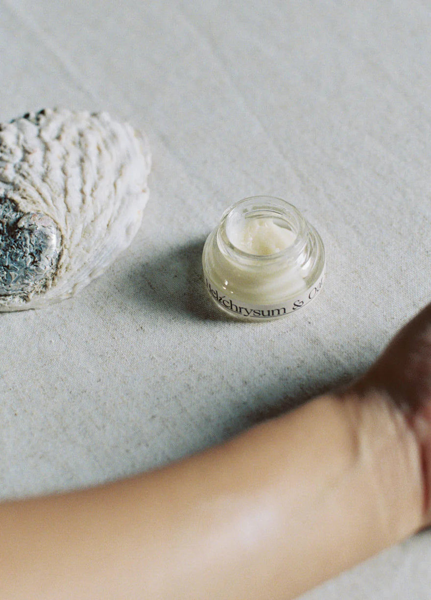 Small glass jar of Palm Of Feronia Helichrysum Oat Cream Glasshouse cream on a textured surface with a hand reaching towards it.