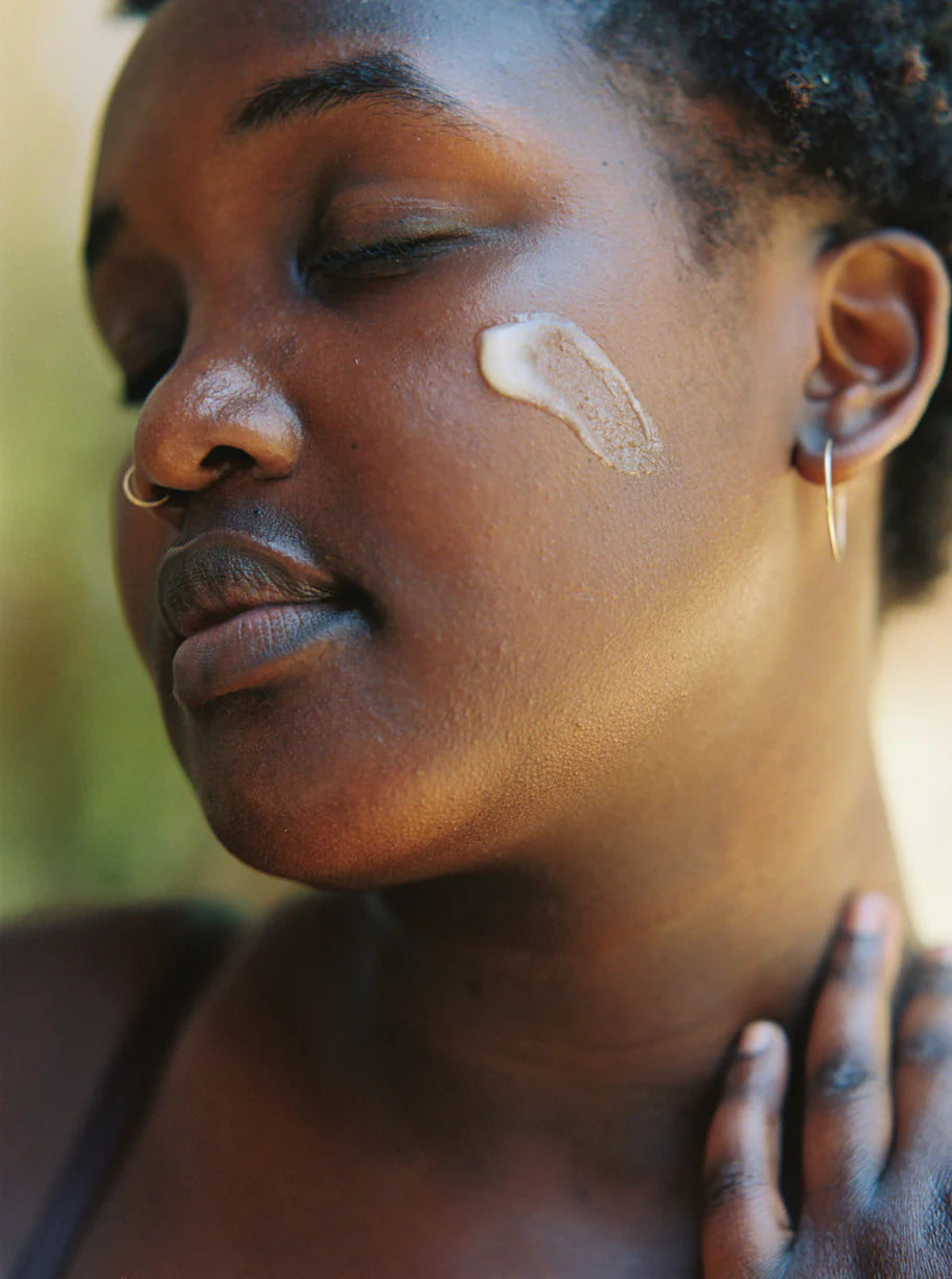 Close-up of a woman's face with a light-coloured product Palm Of Feronia Helichrysum Oat Cream on her cheek, outdoors.
