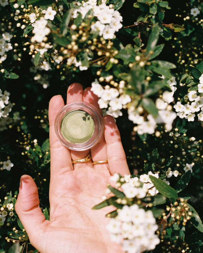 Hand holding a small container of green cream against a natural background with white flowers.