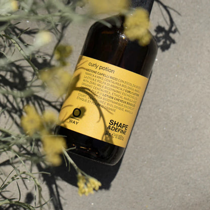 Bottle of 'Curly Potion' by OWAY  a curly hair styling cream with a yellow label on a gray surface with plants.