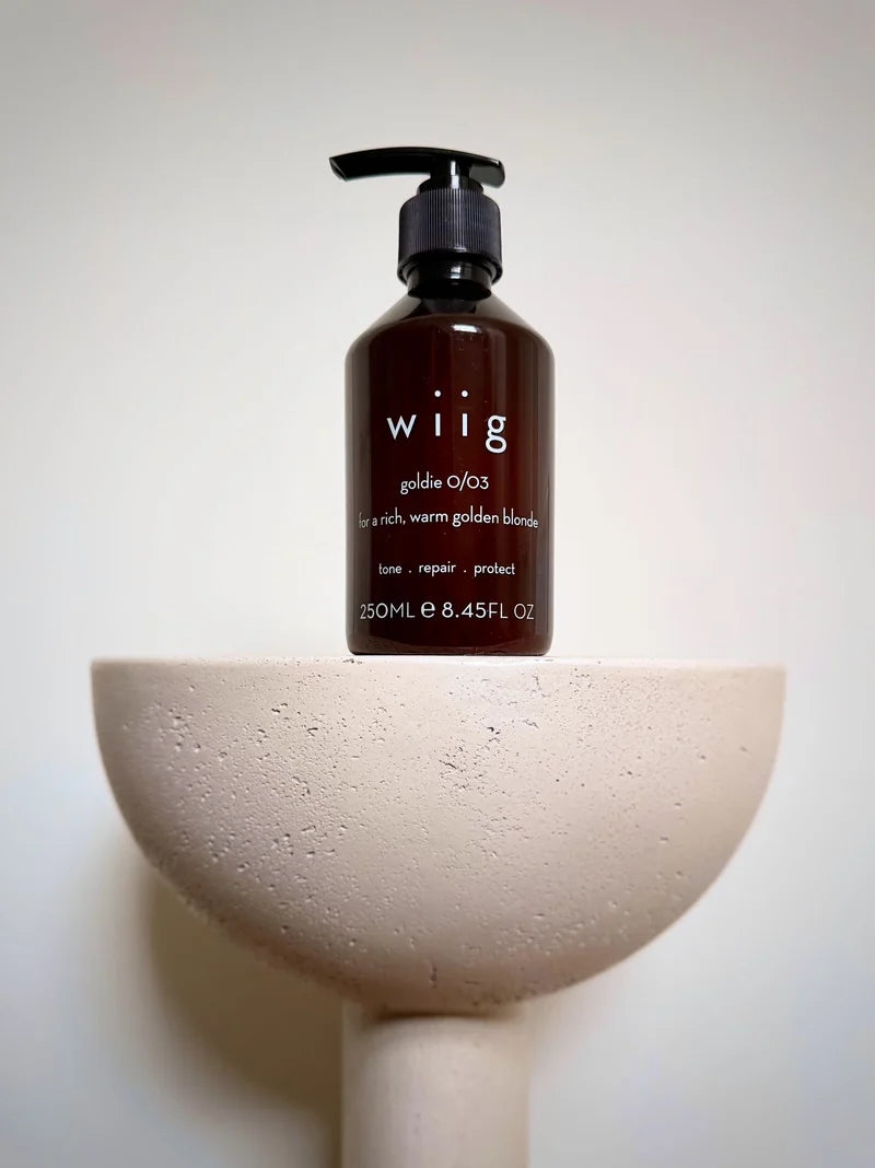 A dark amber pump bottle of wiig hair care product labeled 'golden o/03', displayed on a light beige textured pedestal against a plain off-white background.